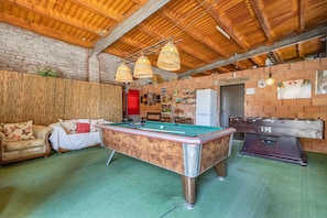 A pool table, sofa, and foosball table in a room with brick walls and wooden beams.
