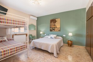 Un dormitori amb lliteres, un llit, dues taules de nit amb làmpades, una cadira i un quadre a la paret.