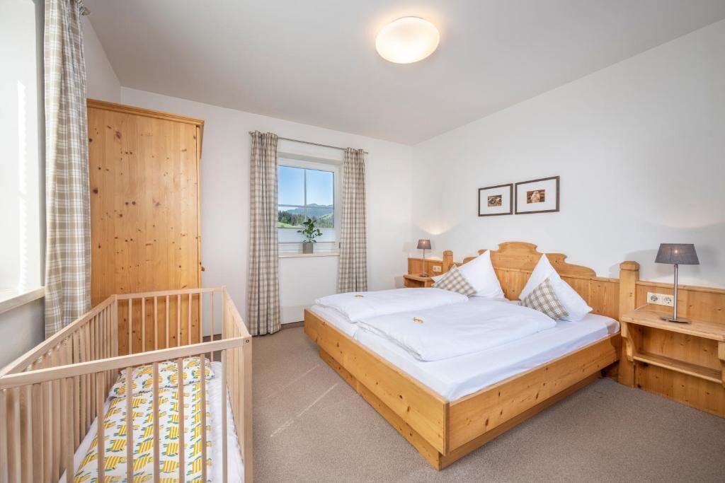 A bedroom with a wooden bed, a crib, a window with curtains, and a nightstand with a lamp.