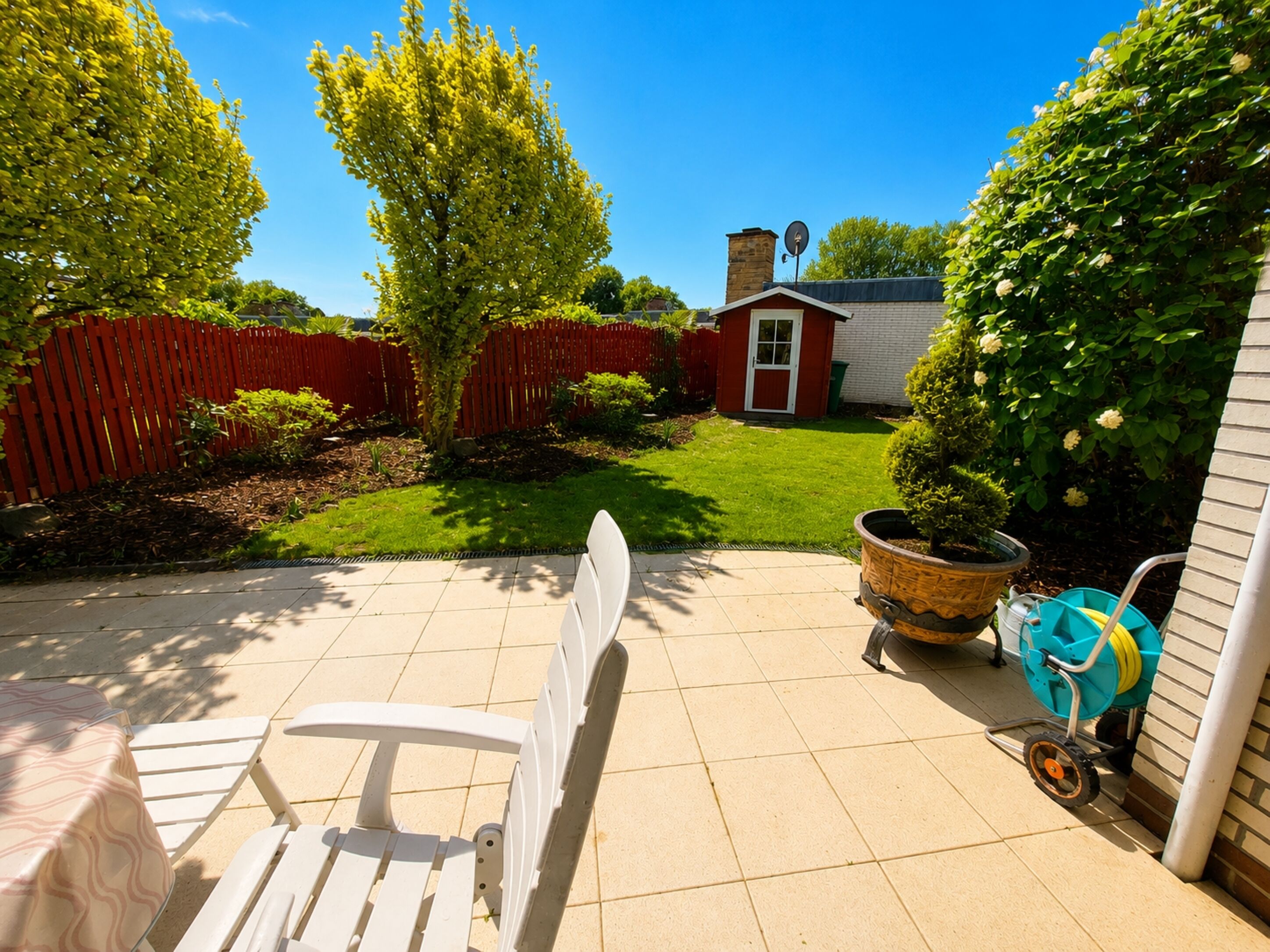 Une terrasse avec une chaise blanche, une brouette et un petit abri de jardin en arrière-plan.