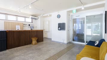 Zona de recepción con un mostrador de madera, un reloj y una puerta de vidrio que da a un área de espera con un sofá amarillo.