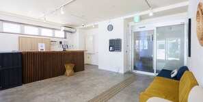 Una zona de recepció amb un taulell de fusta, un rellotge i una porta de vidre que dona a una sala d'espera amb un sofà groc.