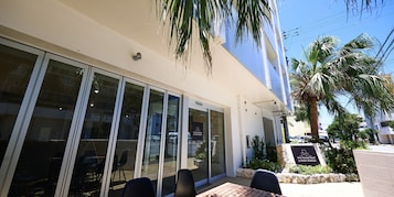 Entrada de un hotel con amplias puertas de vidrio, mobiliario exterior y un letrero con el nombre del hotel.