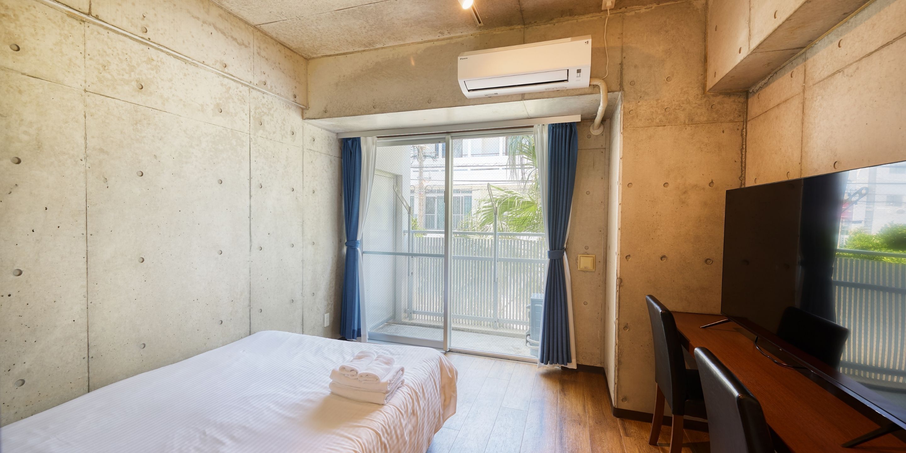 A hotel room with a bed, a desk with a chair, an air conditioning unit, and a view through a sliding glass door.