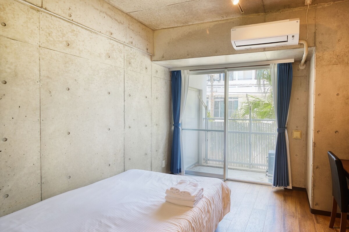A hotel room with a bed, a desk with a chair, an air conditioning unit, and a view through a sliding glass door.