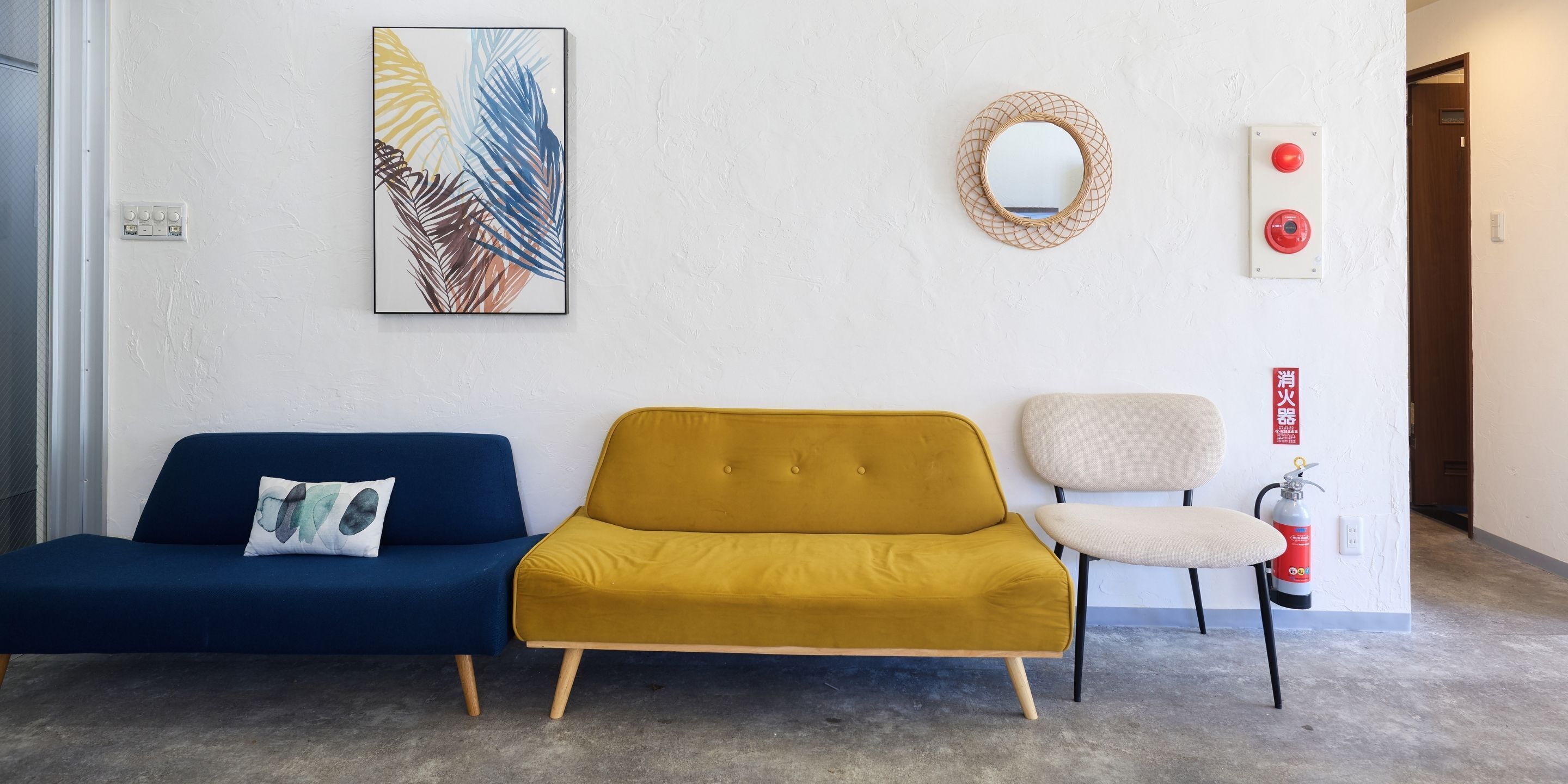A reception area with a blue sofa, a mustard-colored sofa, and a white chair. There is a framed artwork on the wall, a round mirror, and a fire extinguisher.