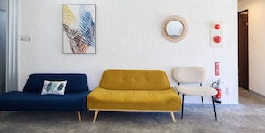 A reception area with a blue sofa, a mustard-colored sofa, and a white chair. There is a framed artwork on the wall, a round mirror, and a fire extinguisher.