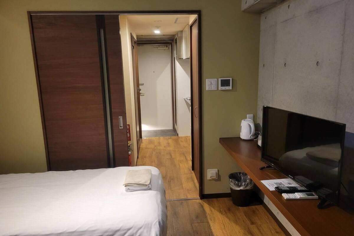 Quarto de hotel com cama, televisão, escrivaninha com computador, lixeira e banheiro visível através de uma porta aberta.