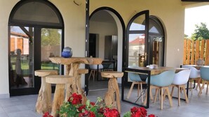 An outdoor patio with a wooden table and chairs, a glass door, and red flowers.
