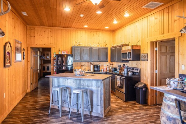 A wooden interior with a kitchen area featuring a central island, bar stools, a refrigerator, a microwave, and a stove.