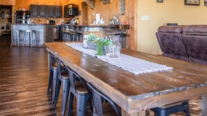 A rustic wooden dining area with a long table, bar stools, and a mounted deer head on the wall.