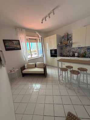 A modern kitchen with white cabinets, a dining area with stools, a sofa, and a window with curtains. - Masticiello House (Nerano)