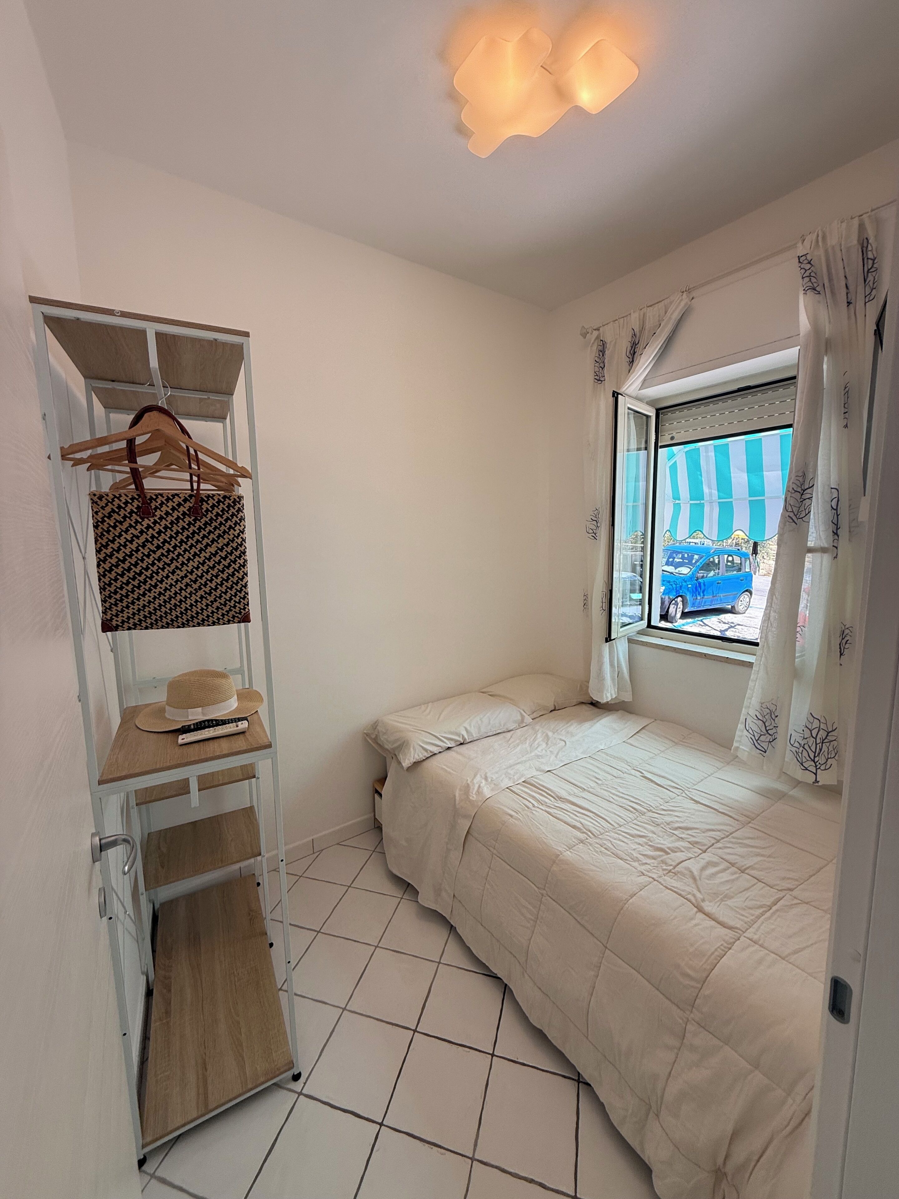 A small, neatly organized room with a bed, a wooden shelf with a hat and remote, a window with curtains, and a view of a blue car outside.