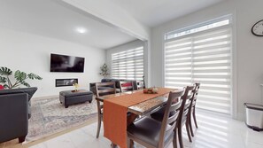 A modern living room with a dining area, a flat-screen TV, and large windows with blinds. - Private 5-bedroom Caledon Home Comfort, Style, and Nature! (Caledon)