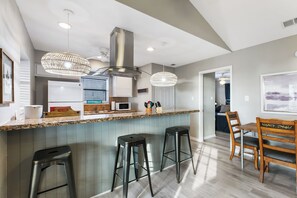 A kitchen with a central island, bar stools, a dining table with chairs, pendant lights, and a stainless steel range hood. - Beachfront South Padre Island condo with bay views, sunsets, and SpaceX launches from your private deck. Sleeps 6 in 2 bedrooms (king and queen) plus convertible sofa. Chef's kitchen, WiFi, washer/dryer, fire pit, grill, free parking. Pet-friendly. (South Padre Island)