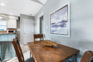 A dining area with a wooden table, chairs, a woven basket, and a framed abstract painting on the wall. - Beachfront South Padre Island condo with bay views, sunsets, and SpaceX launches from your private deck. Sleeps 6 in 2 bedrooms (king and queen) plus convertible sofa. Chef's kitchen, WiFi, washer/dryer, fire pit, grill, free parking. Pet-friendly. (South Padre Island)
