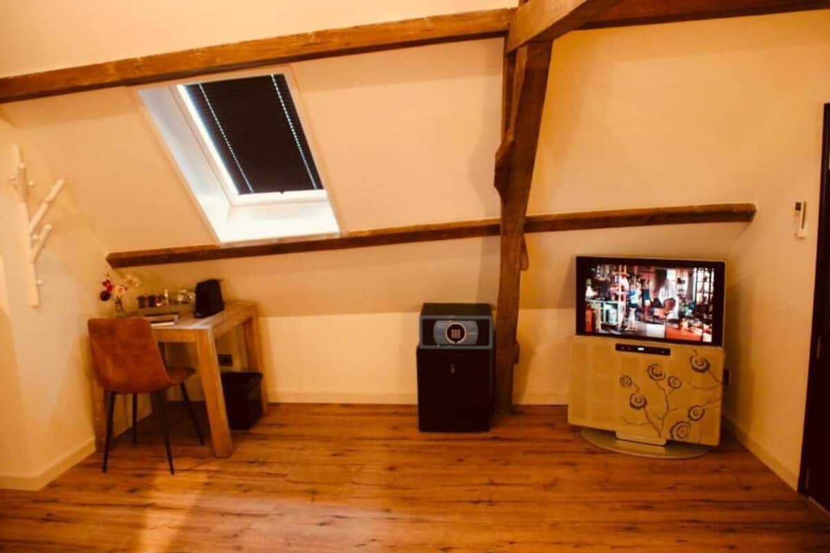 Una habitación con techo inclinado, un ventanal pequeño, un televisor, un escritorio con una silla y un altavoz.