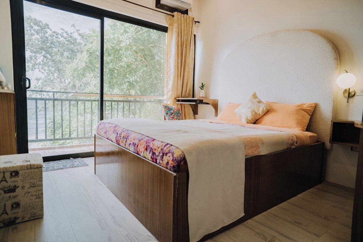A bedroom with a bed, a nightstand, a bedside table, a window with curtains, and a view of trees outside.