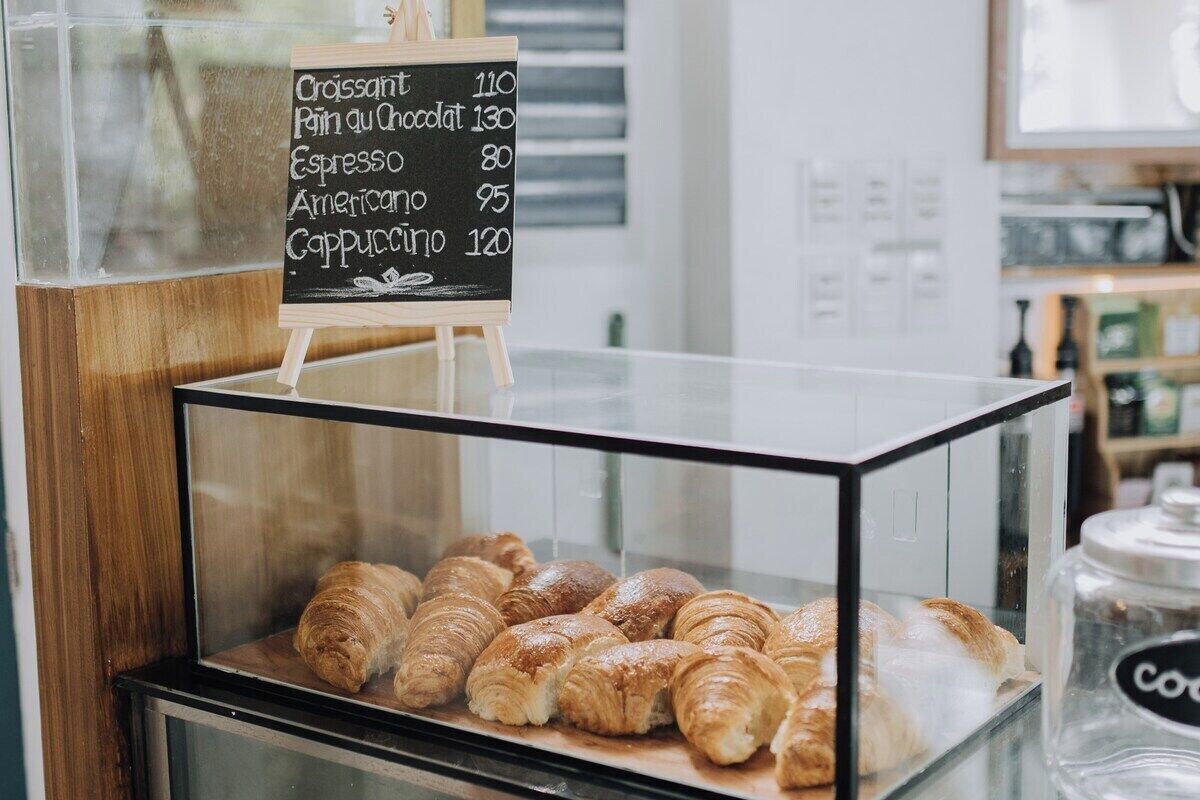 A bakery display case with croissants and a chalkboard menu listing items like croissant, pain au chocolat, espresso, americano, and cappuccino with prices.