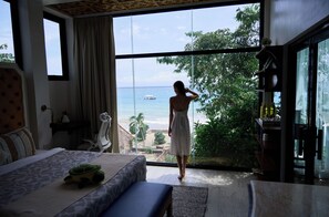A bedroom with a large window, a bed, a bench, and a view of the ocean.