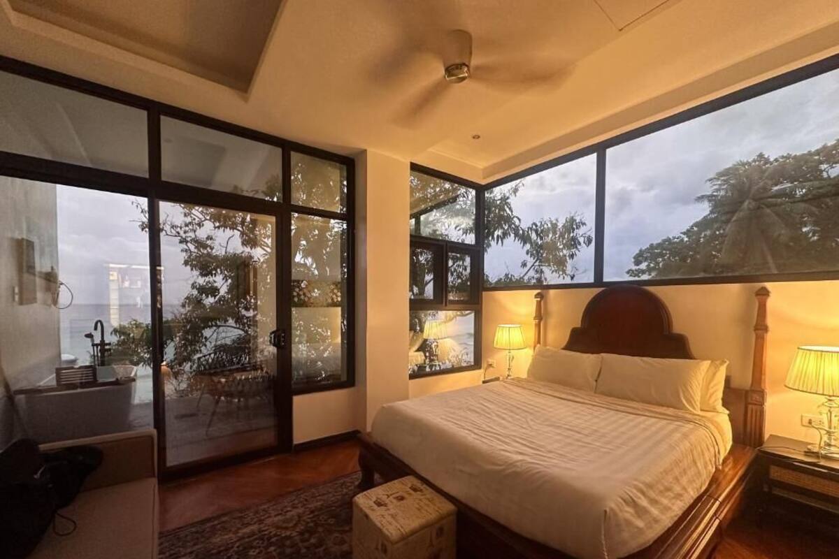 A bedroom with a large bed, a desk, a chair, and a view of trees through the windows.