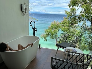 A bathtub with a view of the ocean, a table, and chairs on a balcony.