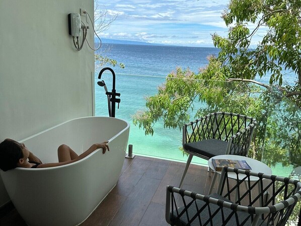 A bathtub with a view of the ocean, a table, and chairs on a balcony.