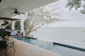 A pool area with a view of the sea, a covered seating area, and a ceiling fan.