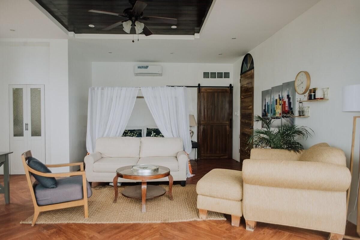 Un salon avec un canapé blanc, une chaise en bois, une table basse ronde, un ventilateur de plafond et une grande fenêtre.