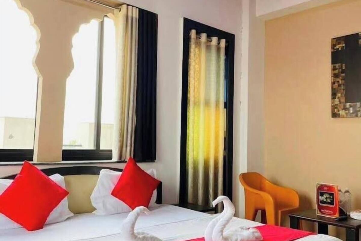 Una habitación de hotel con una cama, almohadones rojos, una decoración de cisne, una ventana, cortinas y una mesita con una botella.