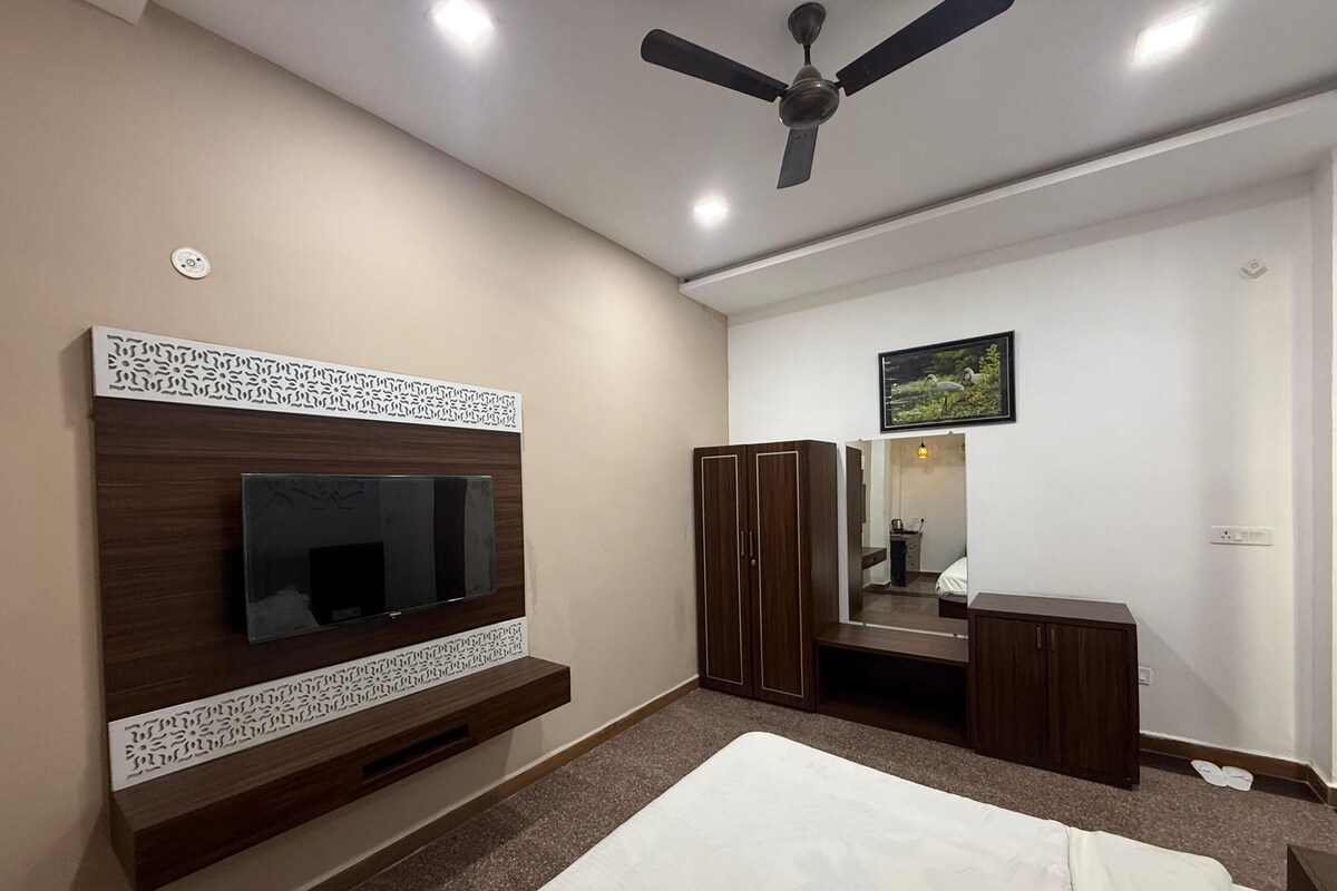 A bedroom with a bed, a TV mounted on a wooden panel, a wardrobe, a mirror, and a ceiling fan.