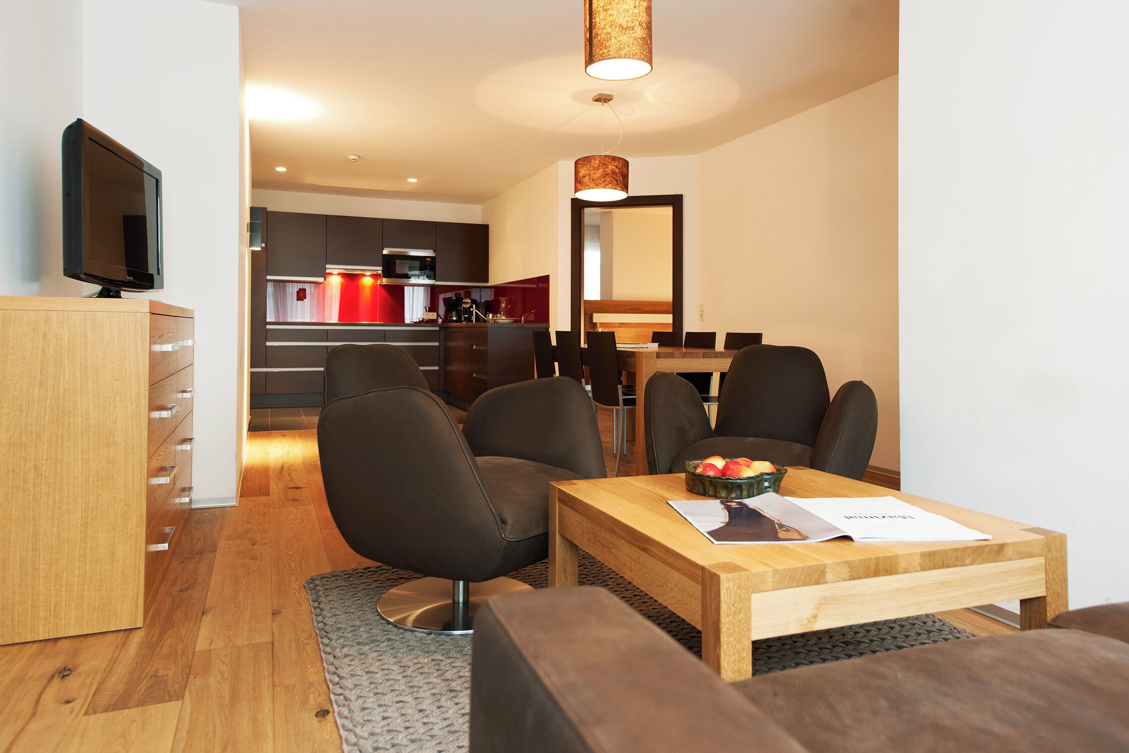 Una sala moderna con una mesa de centro de madera, sillas de cuero negro, un televisor de pantalla plana y una cocina al fondo.