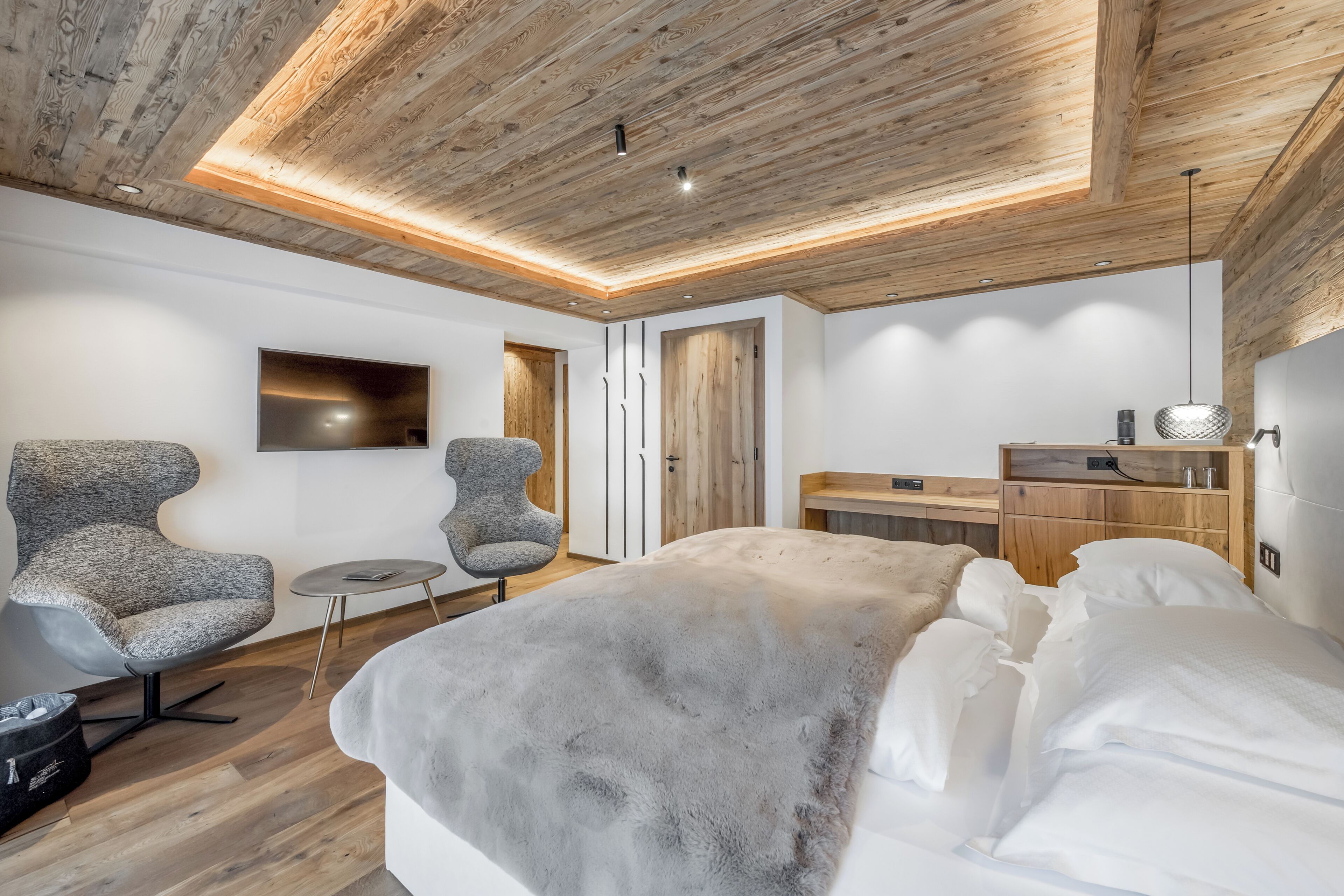 Un dormitorio moderno con techo de madera, una cama grande, dos sillones, una mesita redonda y un televisor montado en la pared.