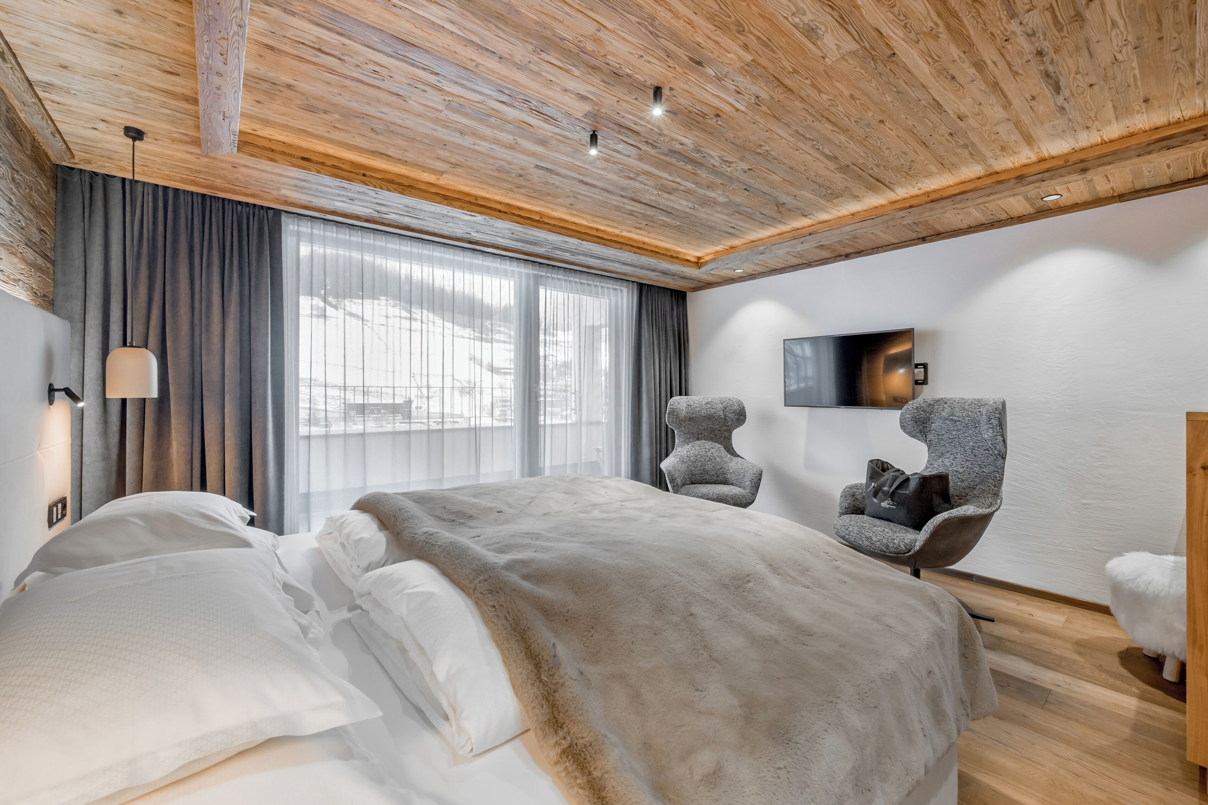 A modern bedroom with a wooden ceiling, a large bed, a chair, a television, and a window with curtains.