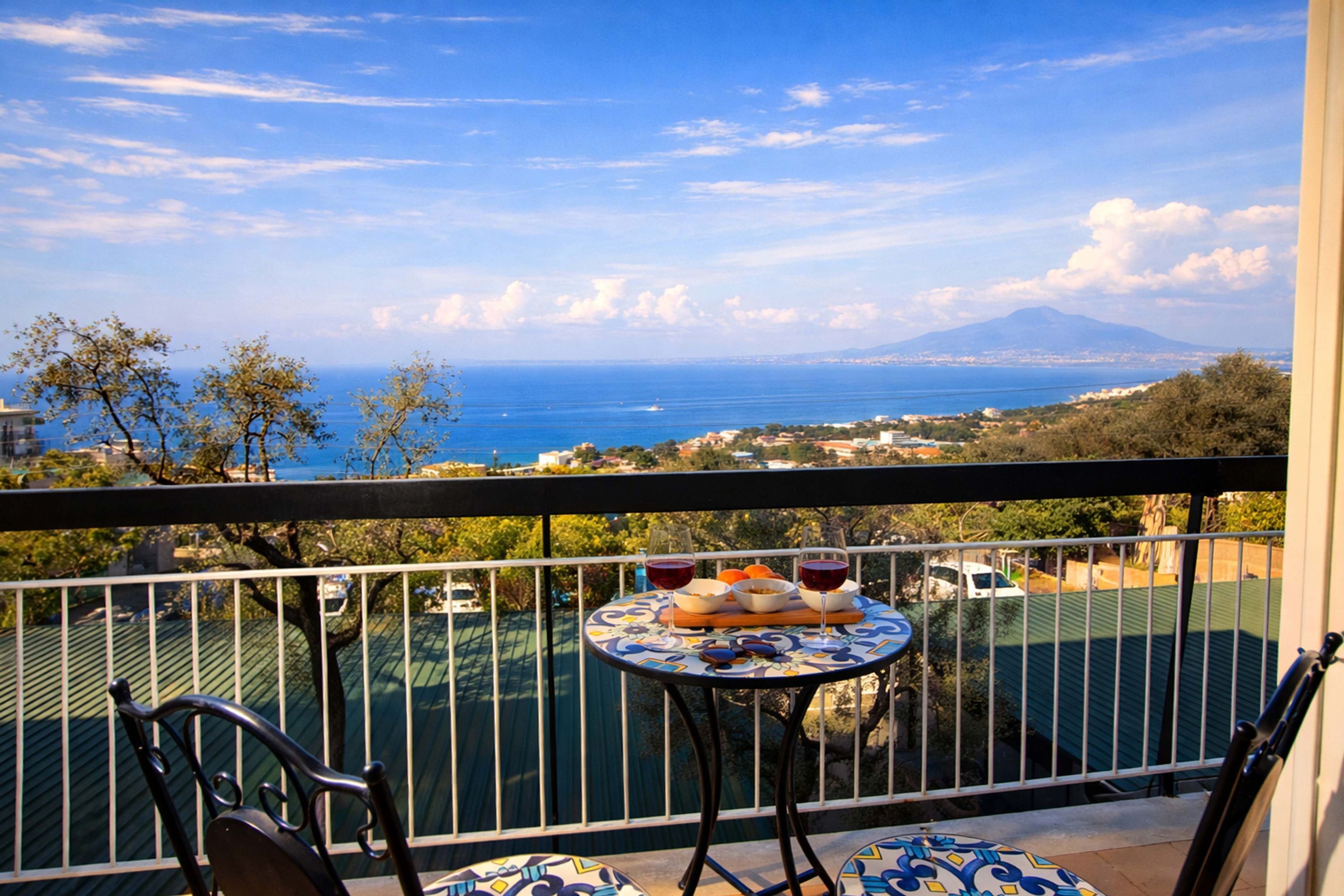 A balcony with a table set for two, overlooking a coastal view with a mountain in the distance.