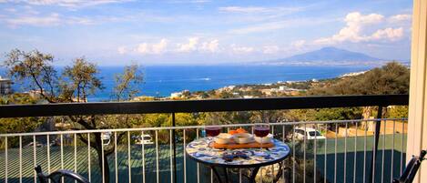 A balcony with a table set for two, overlooking a coastal view with a mountain in the distance.