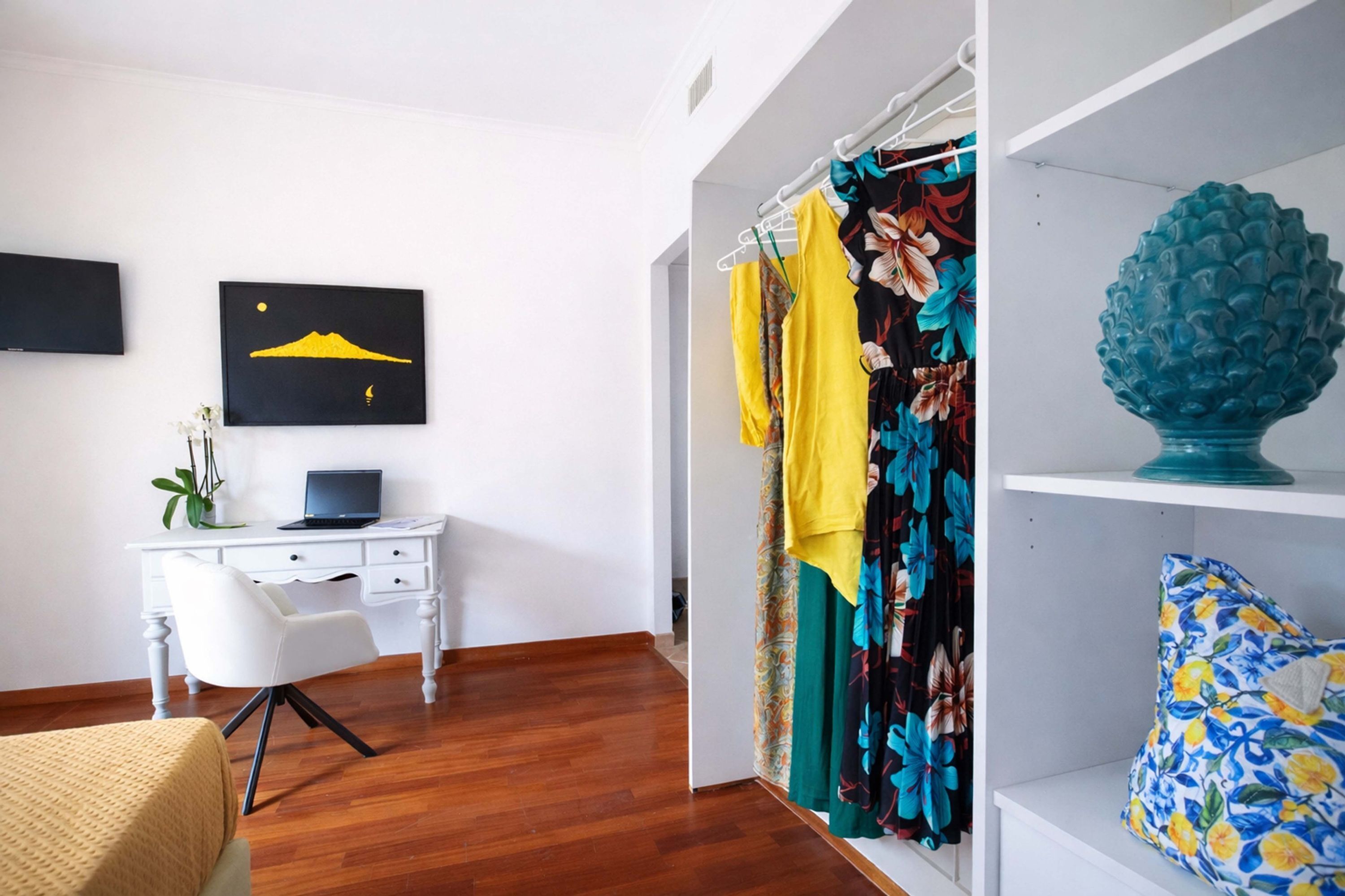 Une pièce avec un bureau blanc, un ordinateur portable, une chaise, un lit, un espace douche agrémenté de rideaux colorés et un vase bleu décoratif posé sur une étagère.