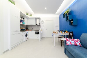 A compact kitchen with a washing machine, a dining table with chairs, and a blue sofa with a floral pillow. - Maison Ann in Sorrento (Sorrento)