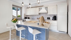 A modern kitchen with white cabinets, a blue island with bar stools, pendant lighting, and a view of the outdoors. - Villa Elios in Massa Lubrense (Massa Lubrense)