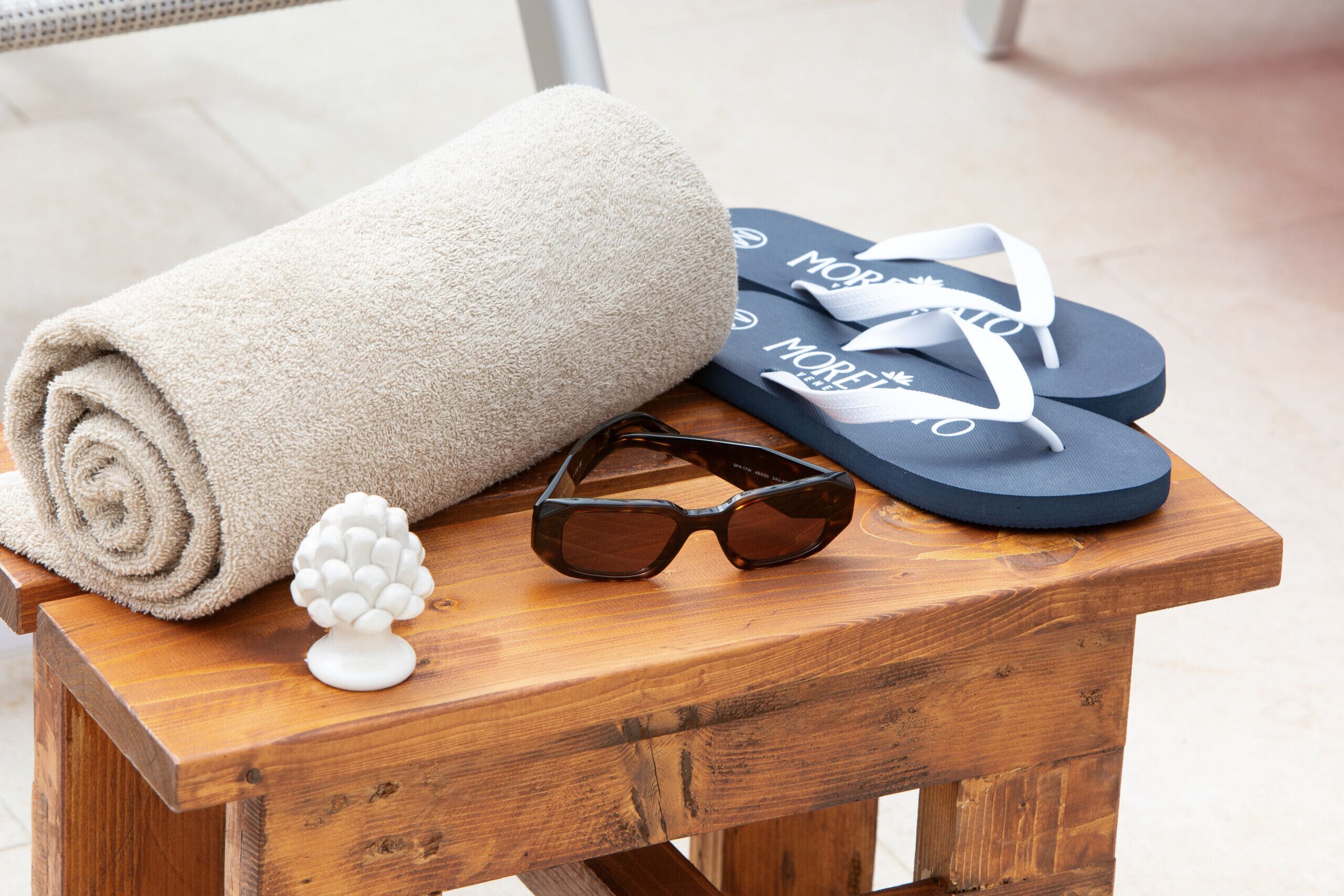 A wooden stool with a rolled beige towel, a pair of black sunglasses, and a pair of blue flip-flops with white text.