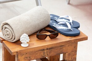 Ein hölzerner Hocker mit einem aufgerollten, beige farbenen Handtuch, einer schwarzen Sonnenbrille und einem blauen Flip-Flop-Paar mit weißer Schrift.