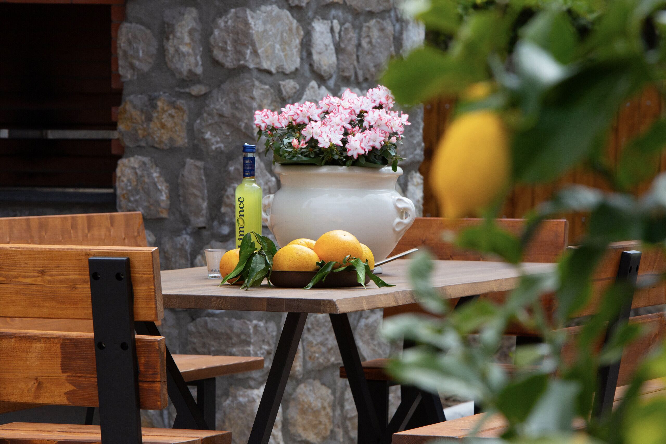 A table with a white vase of pink flowers, lemons, and a bottle of liquor.