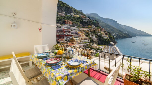 Un balcón con una mesa lista para comer, con vista a un pueblo costero con edificios en una colina.