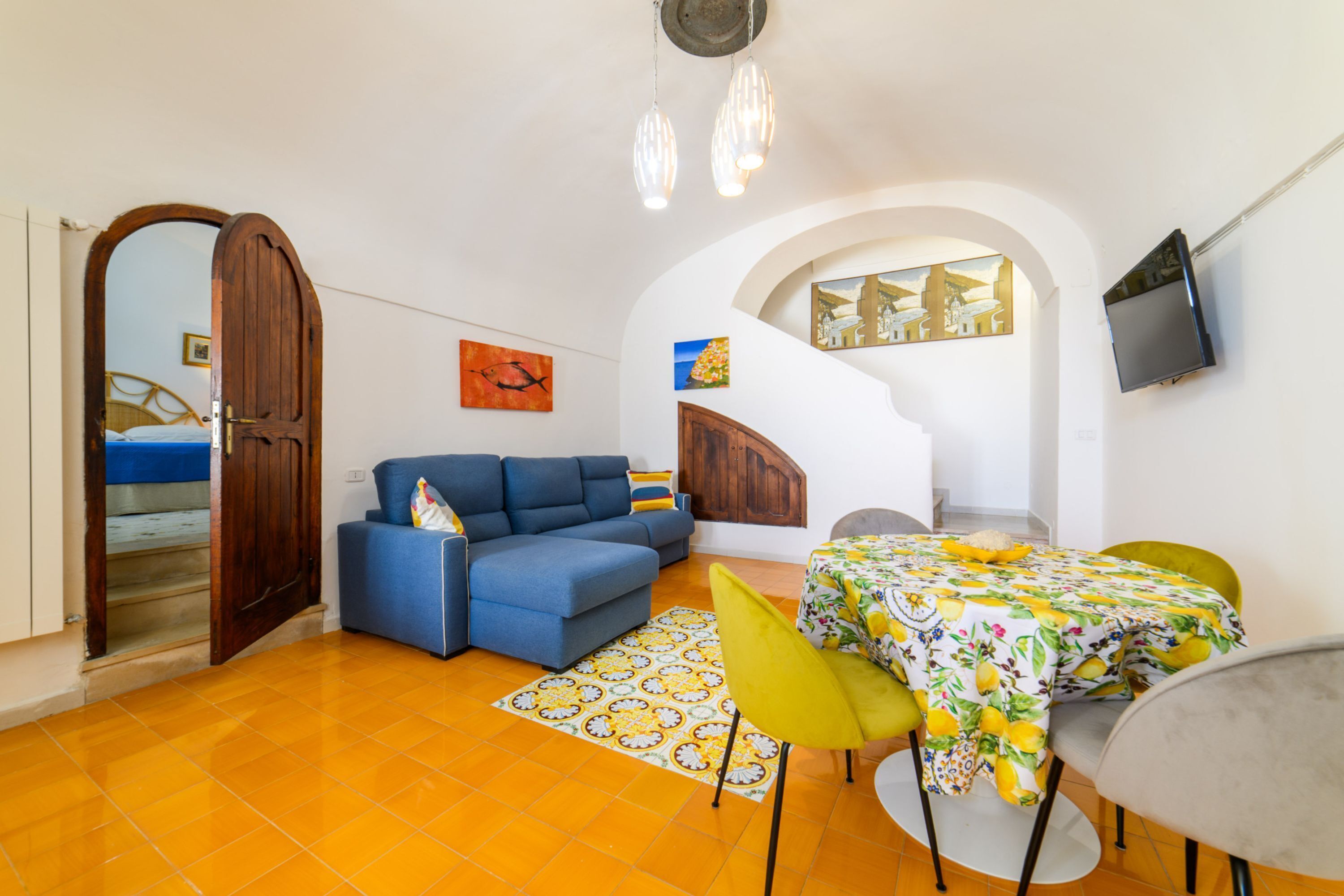 A compact living space with a sofa, dining table, chairs, a TV, artwork, and a staircase.