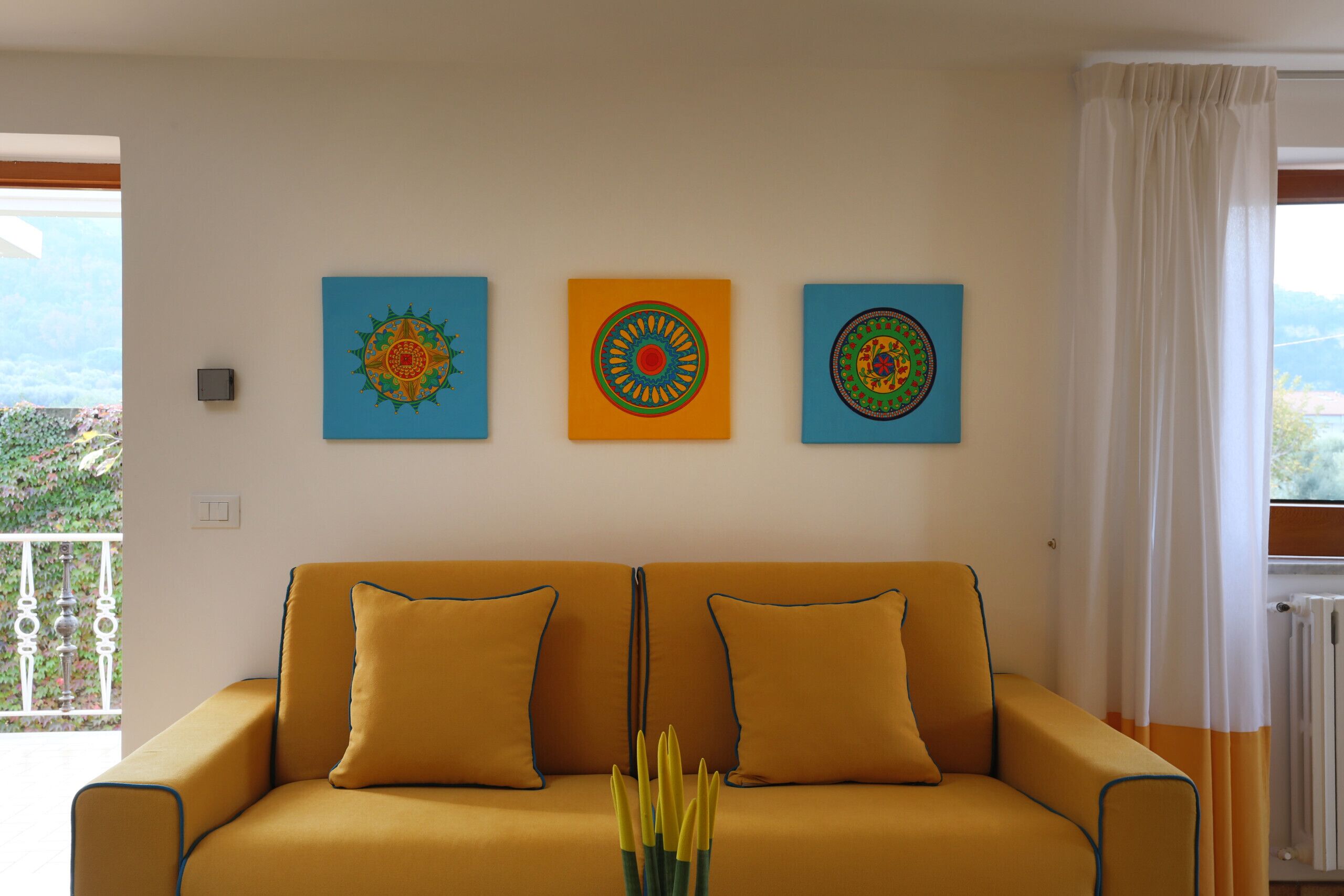 A yellow sofa with two pillows, a wall with three framed artworks, and a view of greenery through a window.