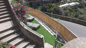 Un balcone con vista su una strada, un recinto in legno e un piccolo giardino con erba sintetica e una panchina.
