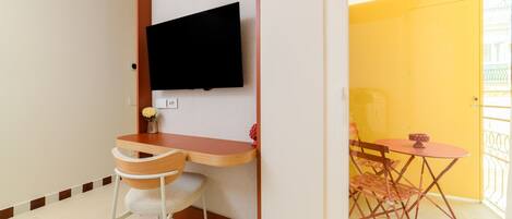 Una habitación con un escritorio de madera, una silla, un televisor montado en la pared y una pequeña mesa con un cuenco.