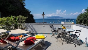 A terrace with lounge chairs, a table, and chairs, overlooking a body of water with boats and a mountain in the distance. - Casa Cinzia - Apartment (Vico Equense)