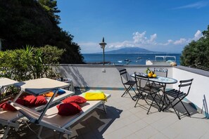 Eine Terrasse mit Liegestühlen, einem Tisch und Stühlen mit Blick auf ein Gewässer, auf dem Boote fahren, und einen Berg in der Ferne.
