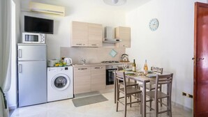 A compact kitchen with a washing machine, microwave, and stove, a dining area with chairs, and a clock on the wall. - Casa Cinzia - Apartment (Vico Equense)